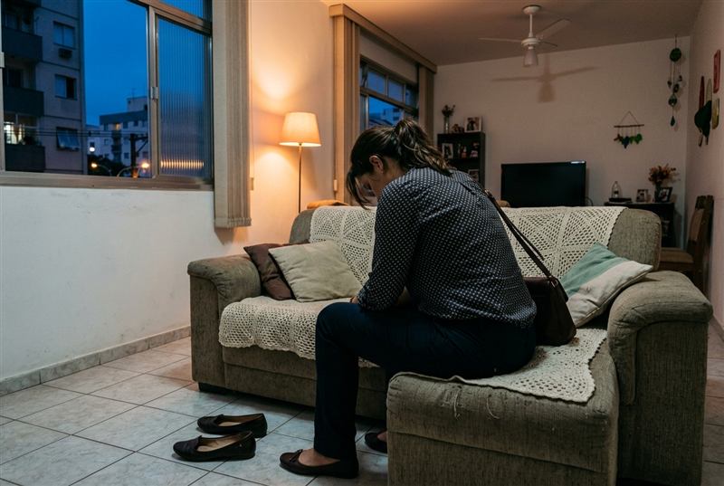 Mulher cansada no sofá no fim do dia, ainda com a bolsa no ombro